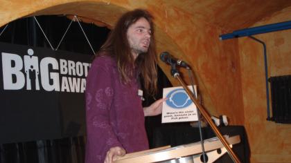 Sebastian Lisken am Redner.innenpult mit einem Schild in der Hand.