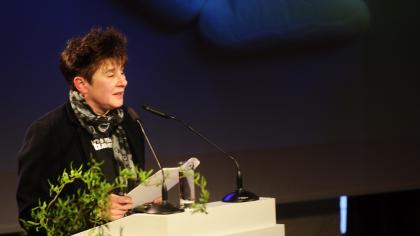 Rena Tangens am Redner.innenpult. Im Hintergrund eine Strichfigur, die auf einer Wolke liegt.